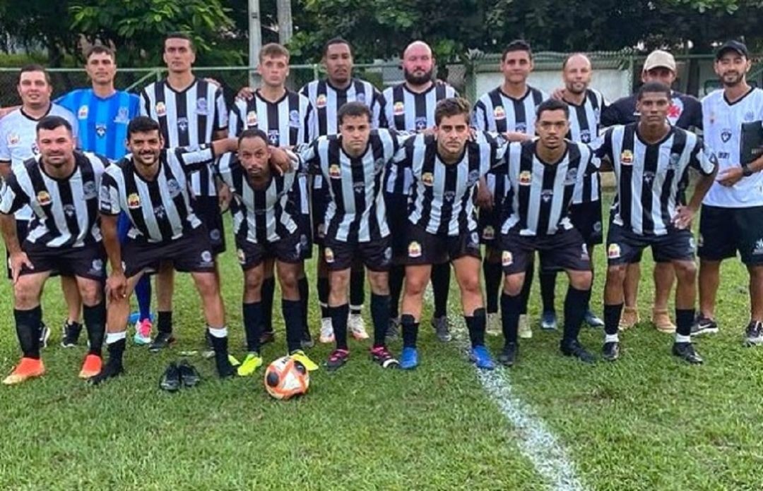Fotografia da equipe masculina de Irupi posada no gramado. Os jogadores vestem uniforme com listras verticais em preto e branco, calções e meiões pretos. Ao fundo, árvores e a rede de proteção do campo de futebol.