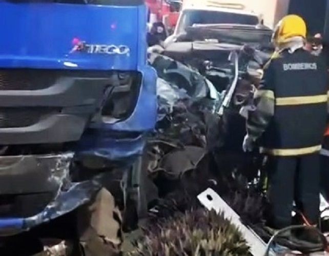 Fotografia em plano fechado da parte frontal de um caminhão azul modelo Atego, que sofreu danos na lateral esquerda após colisão. Ao fundo, destroços de um veículo de passeio totalmente destruído e um bombeiro de costas, usando farda escura e capacete amarelo, avaliando a cena.