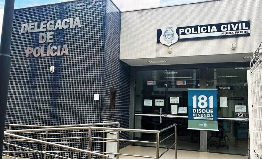 Fachada da Delegacia de Polícia Civil de Muniz Freire, com acabamento em pastilhas escuras, rampa de acesso com corrimão e cartaz do Disque-Denúncia 181 na porta de vidro.