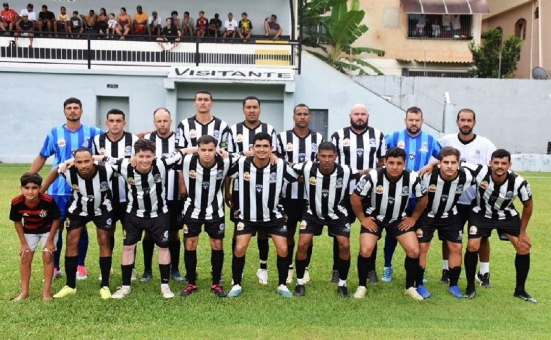 Fotografia oficial da equipe masculina de futebol de Irupi disposta no gramado. Os jogadores vestem uniforme listrado em preto e branco. Onze atletas posam em pé e oito agachados. No canto esquerdo, uma criança com a camisa do Flamengo acompanha o time. Ao fundo, vê-se uma arquibancada com a inscrição 