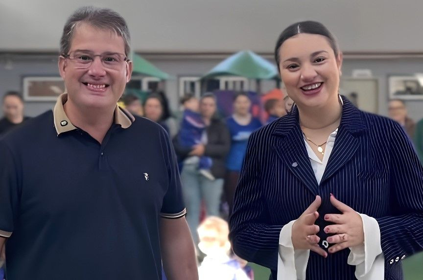 O prefeito Thiaguinho à esquerda, de óculos e camisa polo azul-escura com detalhes em bege na gola. A secretária Esther Simões à direita, com cabelo preso, sorrindo, vestindo blazer azul-escuro risca-de-giz sobre camisa branca e colar dourado, com as mãos articuladas à frente. Ao fundo, de forma desfocada, há uma área externa com brinquedos de playground e pessoas conversando.