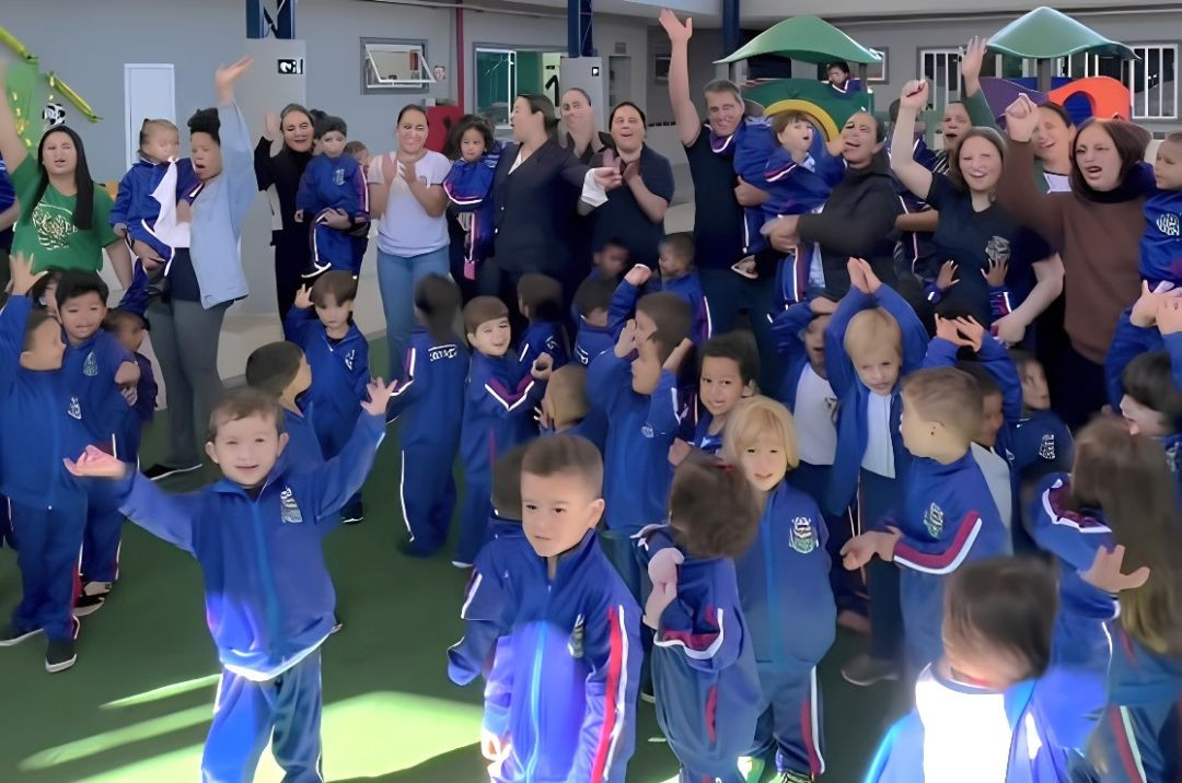 Grupo numeroso de crianças pequenas, alunas da creche municipal, reunidas em um pátio escolar de piso verde. A maioria veste o novo uniforme oficial: um agasalho azul-escuro com faixas vermelhas e brancas nas mangas e o brasão da prefeitura no peito. Algumas crianças e monitoras ao fundo aparecem sorrindo, acenando e aplaudindo em clima de comemoração.