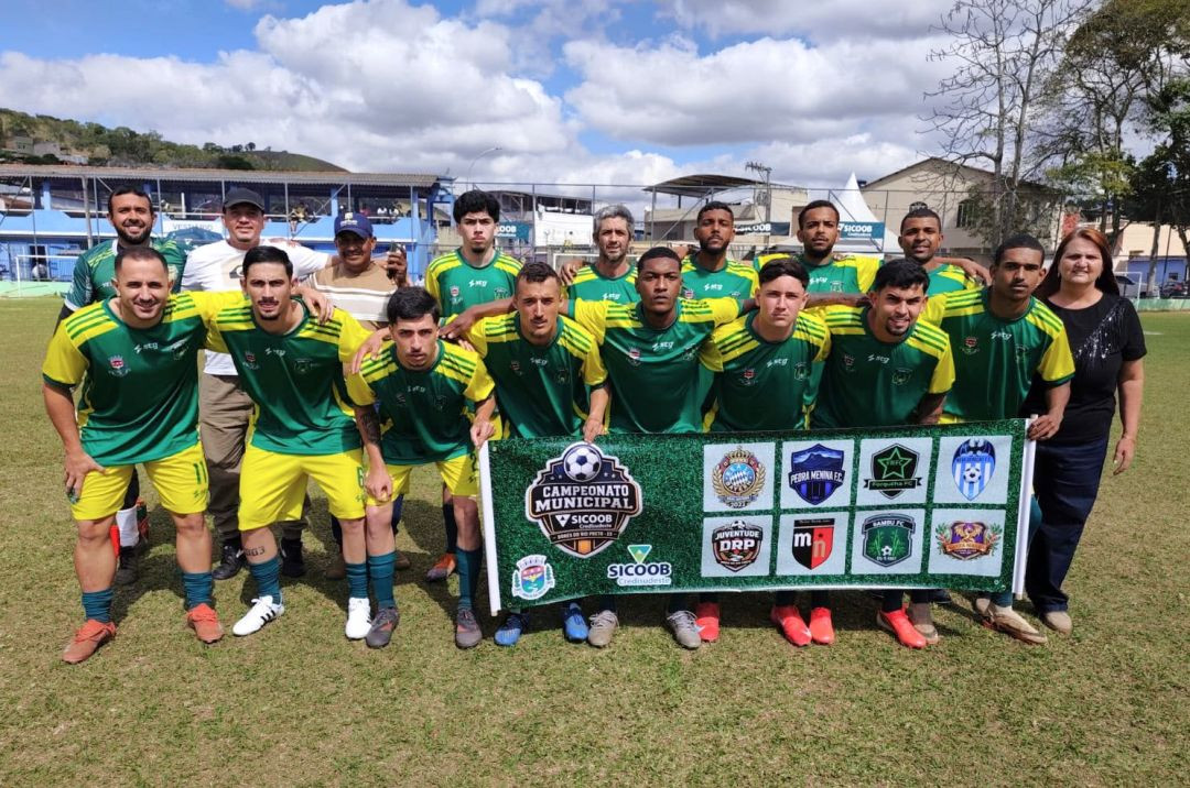 Elenco do Bambu FC em pé e agachado no gramado vestindo uniforme verde e amarelo. Na frente dos jogadores, duas pessoas seguram uma faixa oficial do Campeonato Municipal com os escudos das oito equipes participantes.