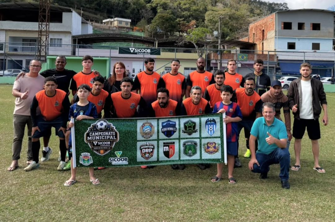 Jogadores do Forquilha do Rio reunidos no gramado vestindo uniforme na cor laranja com detalhes pretos. À frente do grupo, crianças e organizadores seguram a faixa oficial do Campeonato Municipal de Dores do Rio Preto.