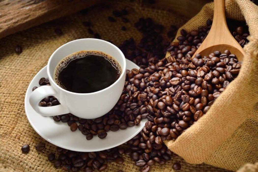 Xícara de café preto sobre um pires cercada por grãos de café espalhados sobre uma superfície de tecido rústico, ao lado de um saco aberto cheio de grãos.