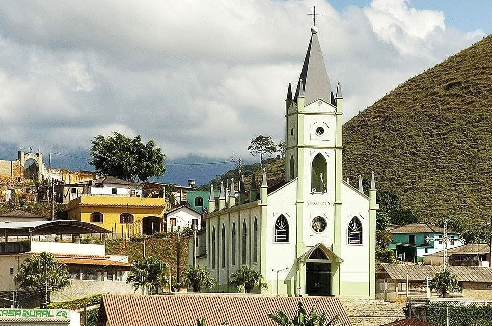 Vista frontal da Igreja Matriz de Divino de São Lourenço, uma construção branca com detalhes em verde claro em estilo neoclássico, situada diante de um morro com vegetação nativa sob céu parcialmente nublado.