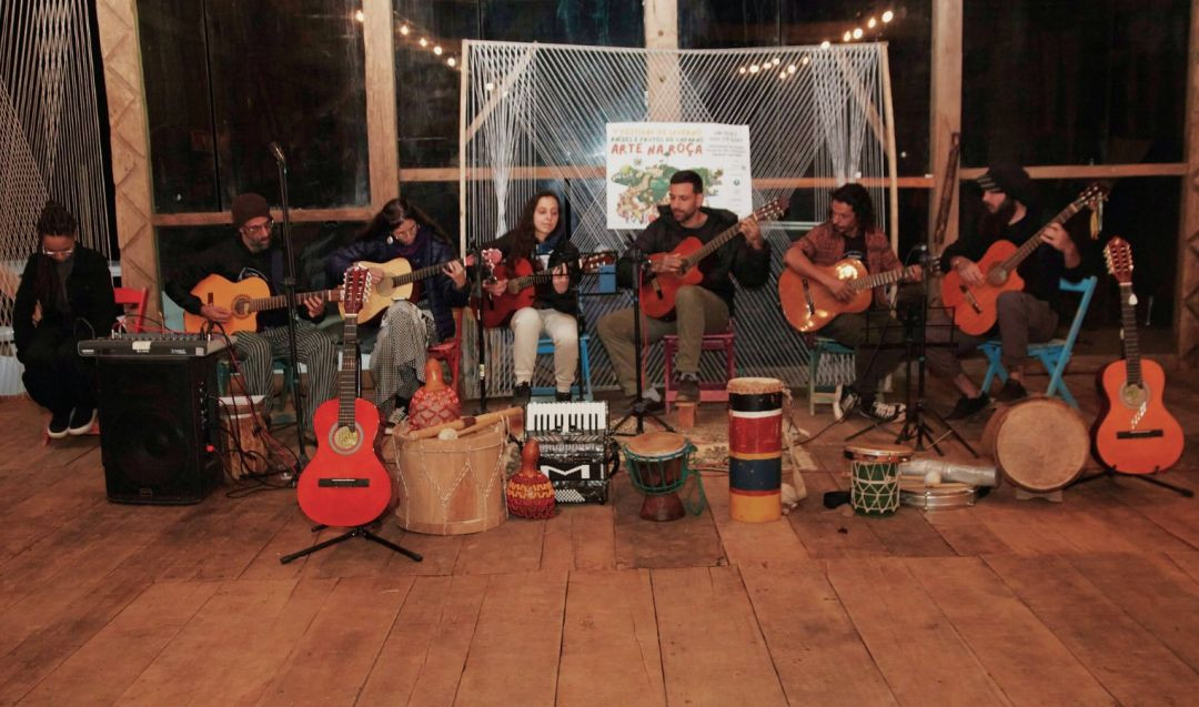 Grupo de seis músicos sentados em cadeiras de madeira tocando violões e instrumentos de percussão em um palco com estrutura de madeira.