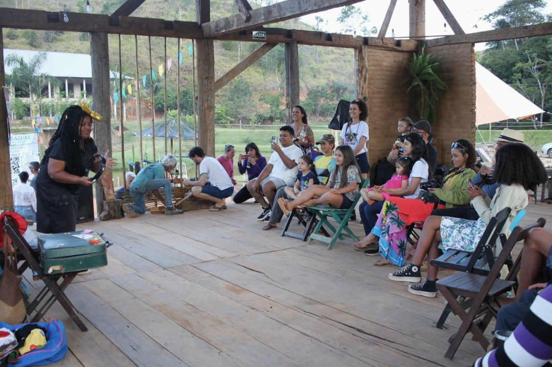 Plateia composta por adultos e crianças assiste a uma apresentação cultural em galpão aberto de madeira e alvenaria.