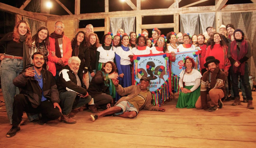 Grupo diversificado de pessoas sorri para a foto segurando estandartes coloridos do grupo de congo no espaço do evento.