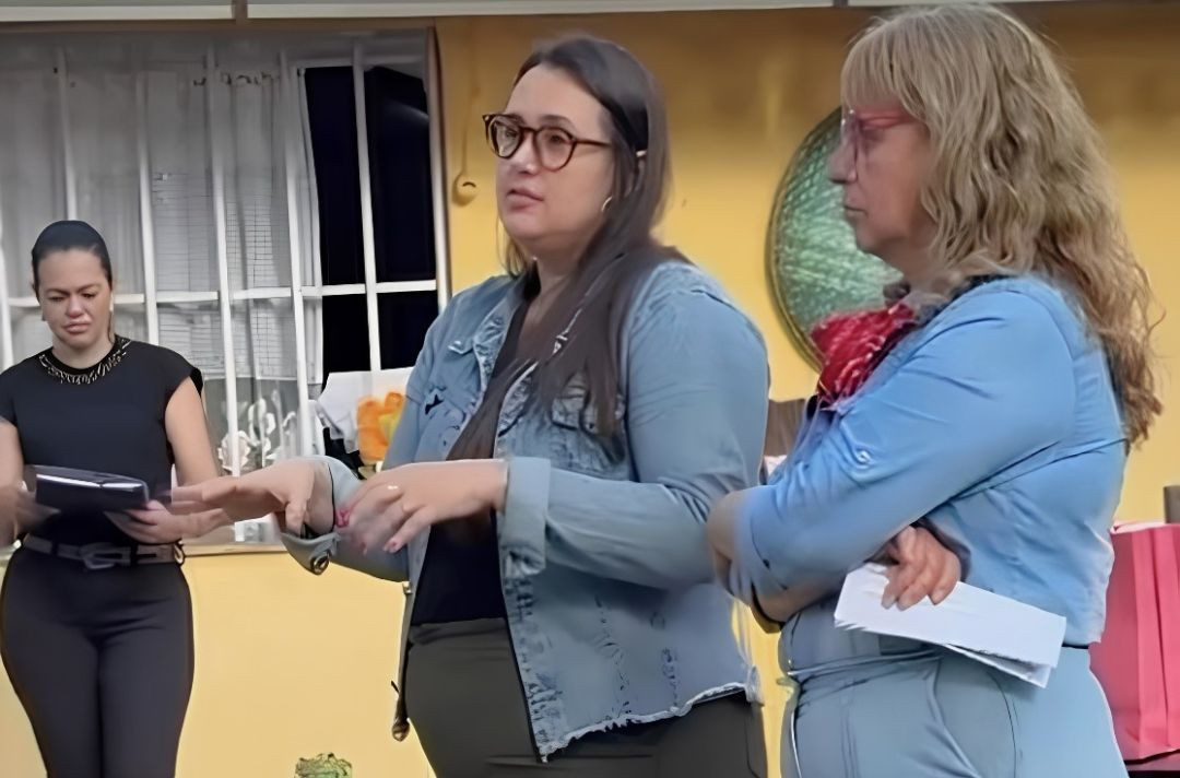 Três mulheres da equipe técnica e gestores, incluindo a secretária Maria Izabel de Souza, em ambiente interno. A mulher ao centro, vestindo jaqueta jeans e óculos, fala e gesticula com as mãos orientando o grupo durante a reunião do programa.