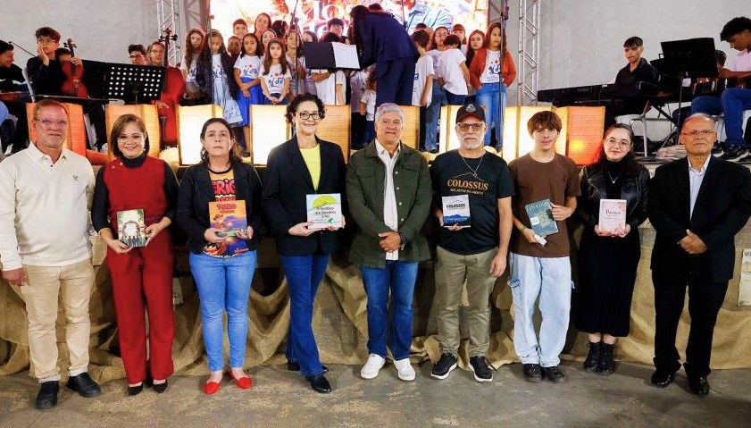 Grupo de homens e mulheres alinhados no palco segurando livros recém-lançados, com crianças do coral posicionadas ao fundo.