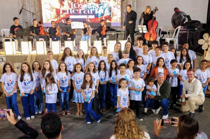 Crianças vestindo camisas brancas do Coral Municipal posicionadas em grupo no palco, sorrindo e acompanhadas pelos professores e autoridades.