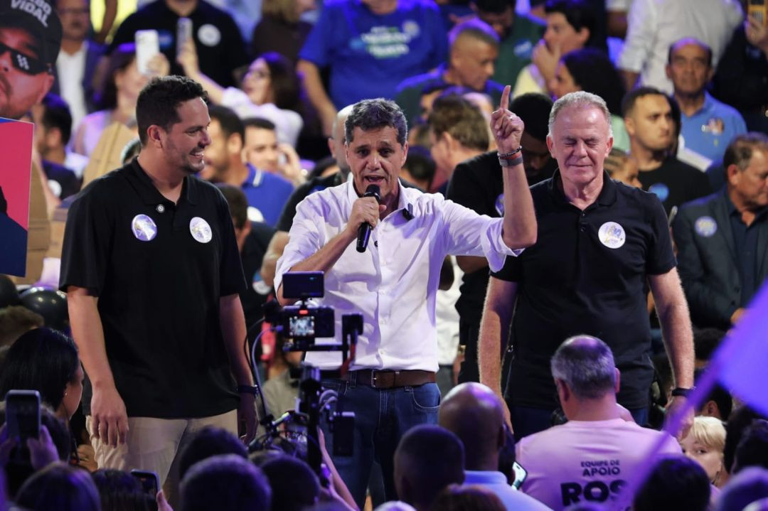 Ricardo Ferraço no centro do palco, usando camisa branca e calça jeans, fala ao microfone e aponta o indicador para cima. Ele está acompanhado do ex-governador Renato Casagrand e o candidato a vice, Camilo Neves, vestidos com camisas polo pretas.