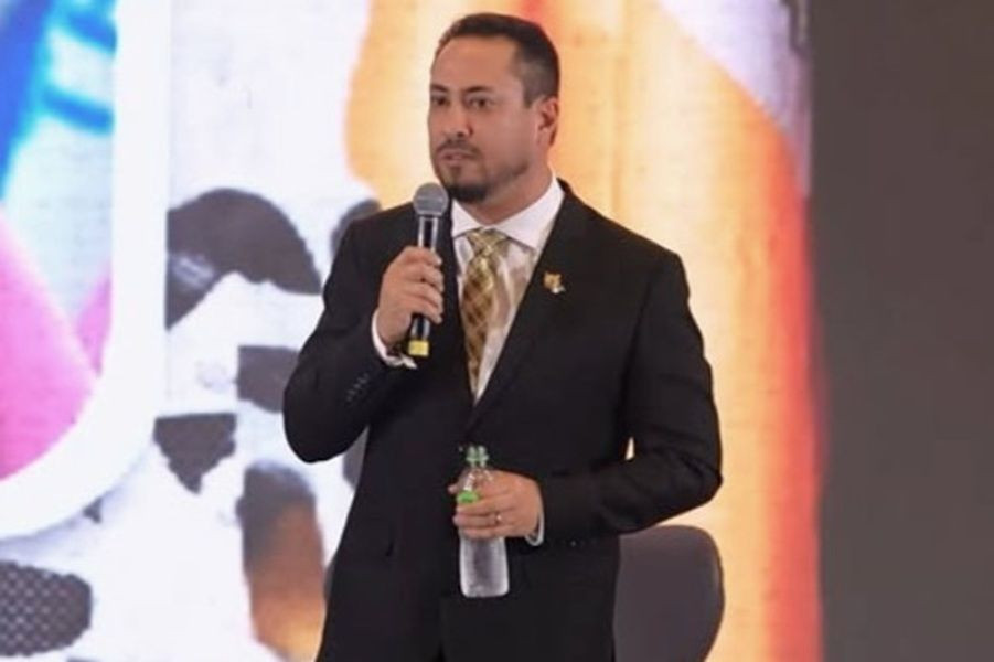 Breno Barcelos de terno escuro, gravata amarela estampada e barba aparada, segurando um microfone na mão direita e uma garrafa de água na mão esquerda enquanto discursa.