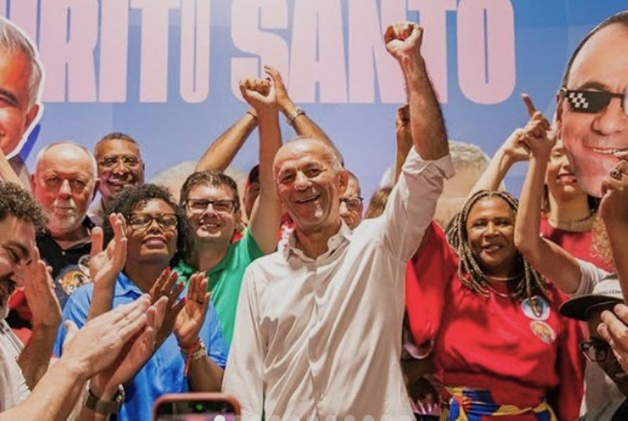 Helder Salomão sorrindo com o punho esquerdo erguido para o alto, vestindo camisa social branca, cercado por apoiadores batendo palmas em frente a um painel com a palavra 'Espírito Santo'.