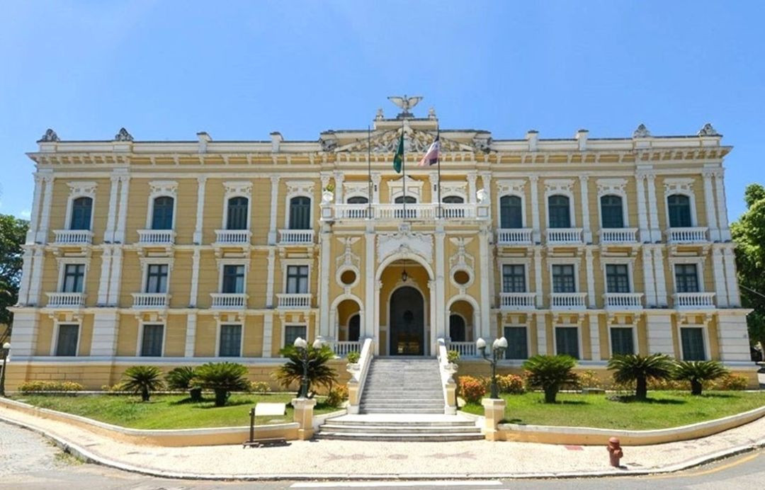Fachada do Palácio Anchieta, edifício histórico amarelado com detalhes arquitetônicos em branco e escadaria central, sob céu azul.