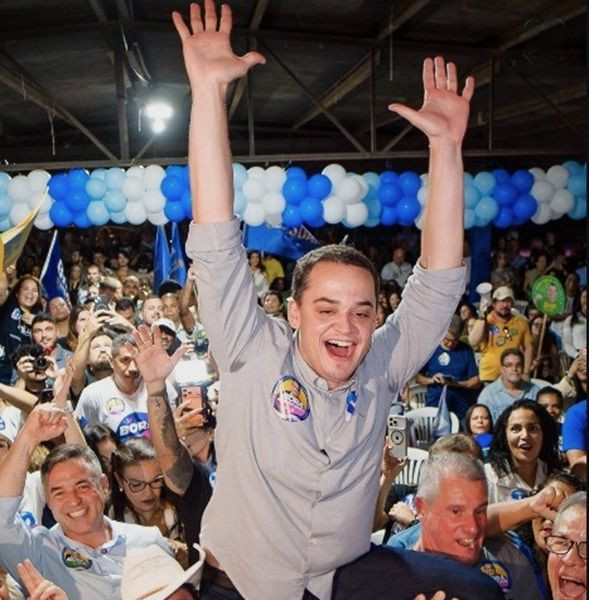 Lorenzo Pazolini com os braços erguidos com as mãos abertas, vestindo camisa cinza e adesivo no peito, sendo carregado nos ombros em meio a uma multidão com balões azuis e brancos ao fundo.