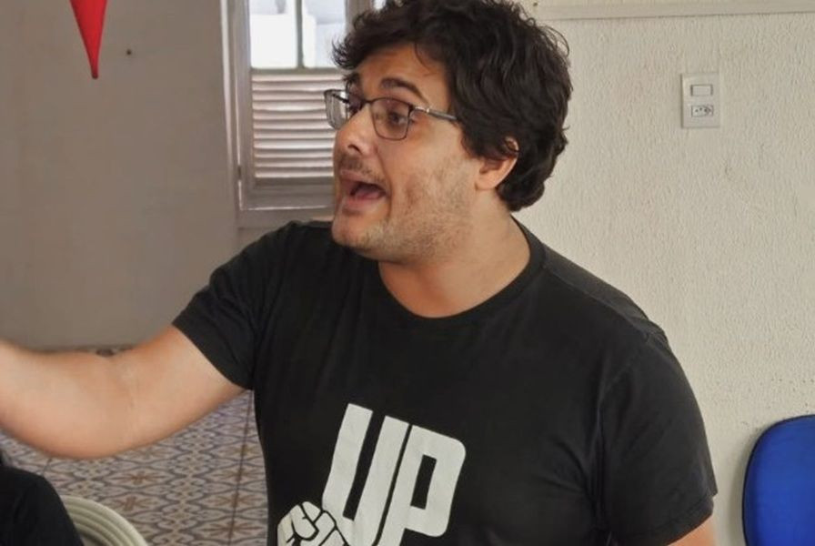 Rafael Demuner de óculos e cabelo escuro gesticulando enquanto fala, usando uma camiseta preta com a sigla 'UP' e o desenho de um punho fechado estampado em branco.