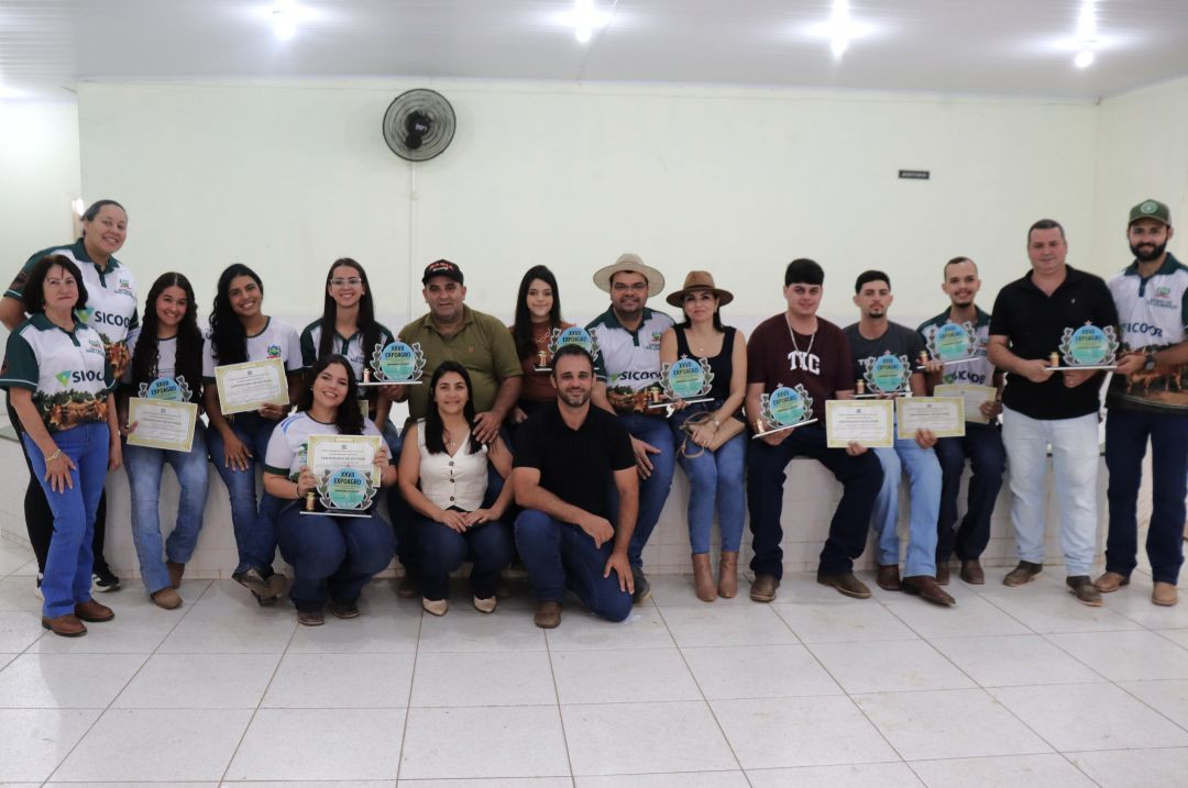 Grupo de produtores rurais posa sentado e em pé segurando certificados e troféus em sala com paredes claras durante a premiação do concurso leiteiro.