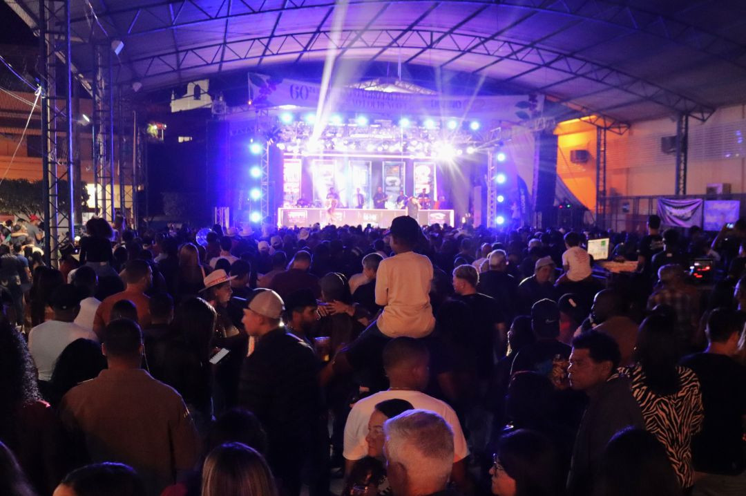Visão posterior de grande público reunido à noite em área coberta iluminada por refletores azuis em frente ao palco do evento.