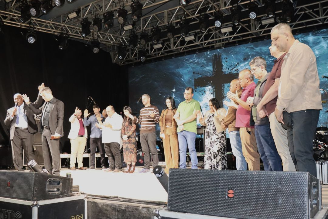 Homens e mulheres alinhados no palco com mãos postas ou erguidas durante celebração de abertura do evento.