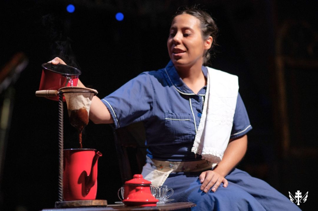 Atriz em cena de espetáculo teatral servindo café coado em um bule vermelho sobre a mesa.