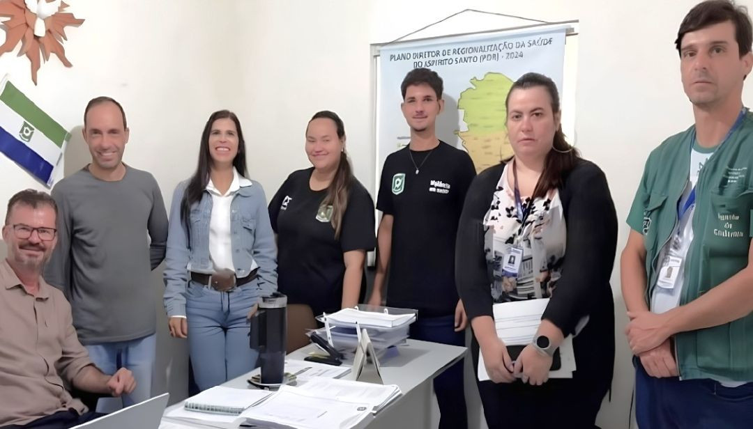 Grupo de profissionais da área da saúde reunido em uma sala de Ibitirama, com documentos sobre uma mesa e um mapa do Espírito Santo ao fundo.
