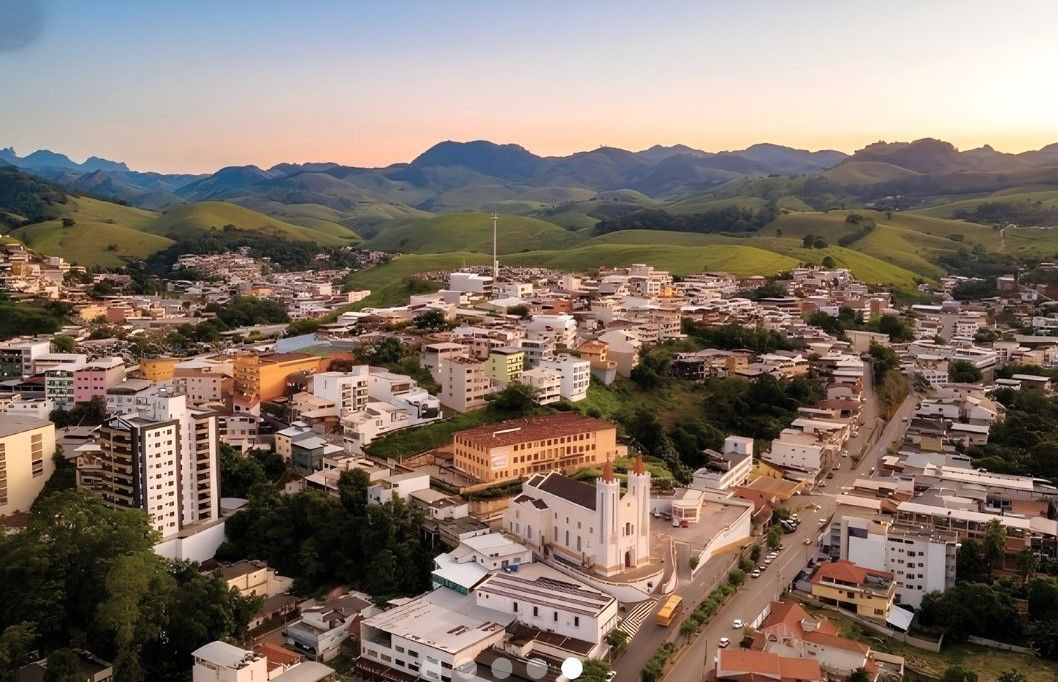 Fotografia aérea da cidade de Alegre mostrando a área urbana com morros verdes e montanhas ao fundo durante o pôr do sol.
