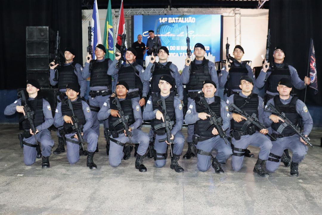 Quatorze policiais militares, vestindo fardas operacionais cinzas, boinas pretas e coletes balísticos, posam para foto oficial no Centro Multiuso de Iúna. Sete estão ajoelhados à frente e oito em pé atrás. Todos seguram fuzis. Ao fundo, um telão azul com o texto 