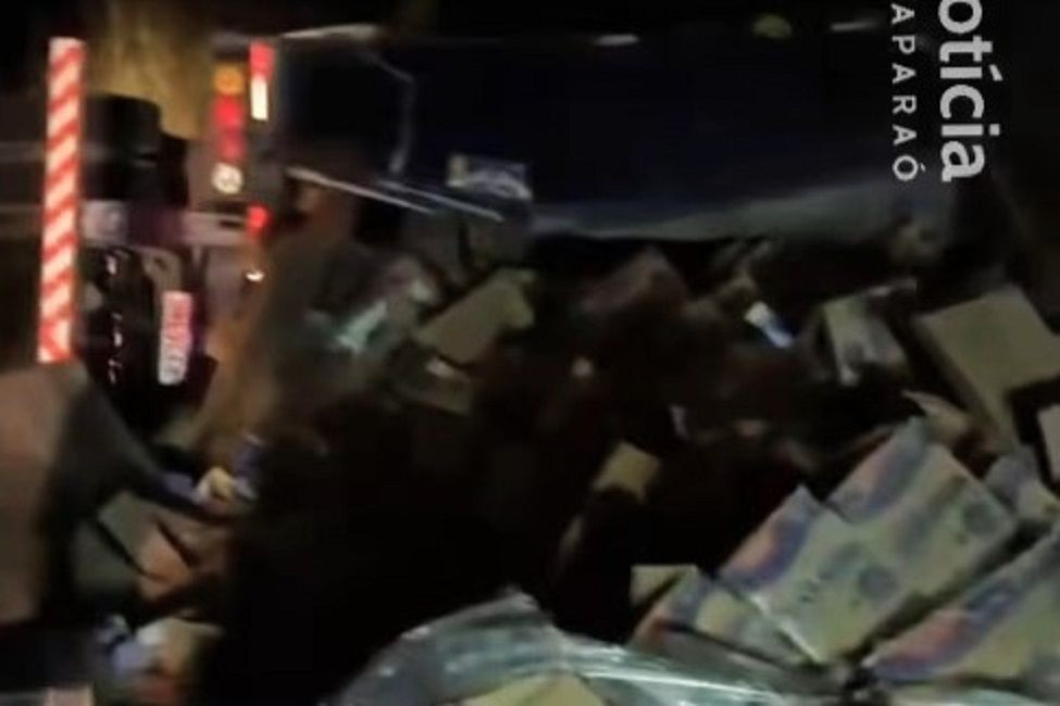 Foto noturna mostrando a traseira e a lateral de um caminhão-baú tombado na pista, com várias caixas de sabão em pó espalhadas no chão ao seu redor.