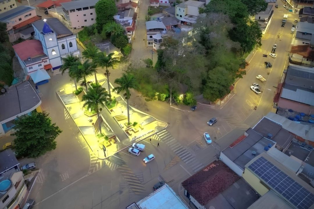 Vista aérea noturna e iluminada do centro de Dores do Rio Preto, mostrando a praça principal, cruzamentos, ruas asfaltadas e construções ao redor.