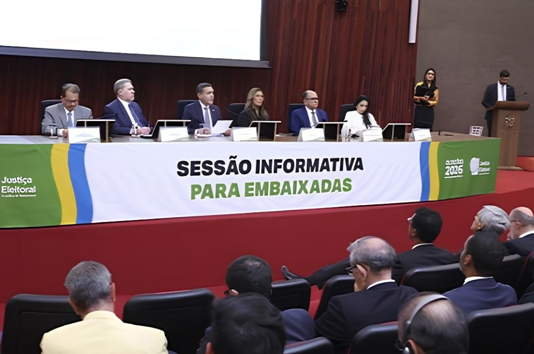 Fotografia do plenário do TSE durante a Sessão Informativa para Embaixadas. No topo, a mesa de honra composta por autoridades e ministros sentados diante de um painel branco e verde. Em primeiro plano, a plateia vista de costas, acompanhando a exposição nas poltronas do auditório.