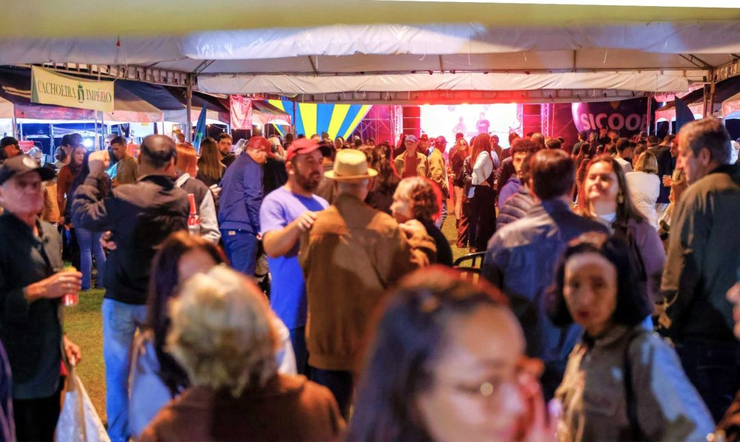 Vista interna do galpão coberto do Festival Gastronômico de Rio Claro com público circulando entre as barracas e a estrutura de luzes do palco ao fundo.