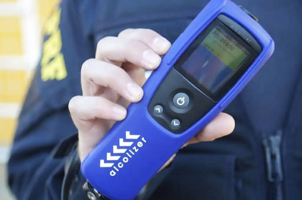 Agente de trânsito vestindo farda escura segura com a mão um etilômetro portátil de cor azul com a tela digital acesa.