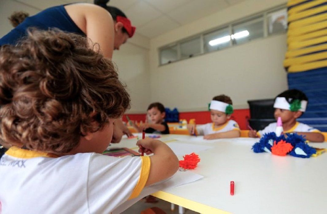 Crianças sentadas em volta de uma mesa branca em sala de aula, desenhando com lápis de cor e manipulando materiais artísticos sob a supervisão de uma professora.