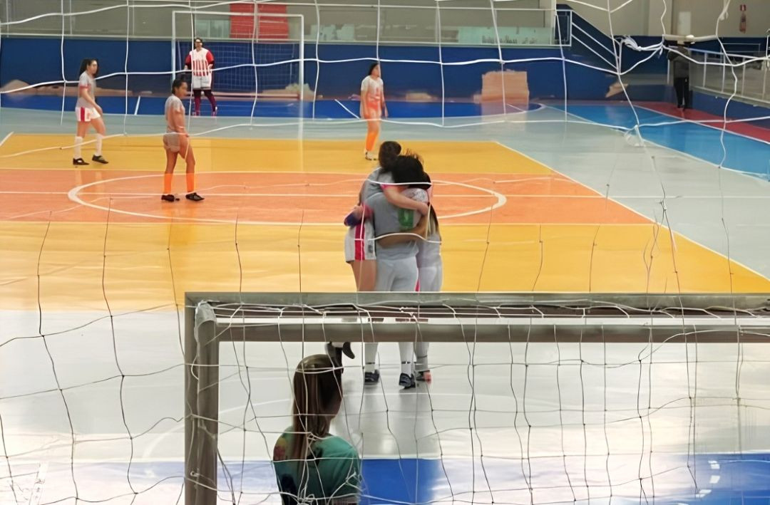 Jogadoras de futsal feminino vestindo uniforme cinza abraçadas em comemoração no centro da quadra, consolidadas por trás da rede do gol.