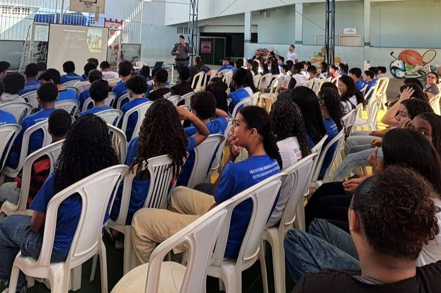 Vista da quadra coberta de uma escola com alunos uniformizados sentados em cadeiras brancas de plástico assistindo a uma palestra apresentada por um policial militar ao fundo, próximo a um telão.