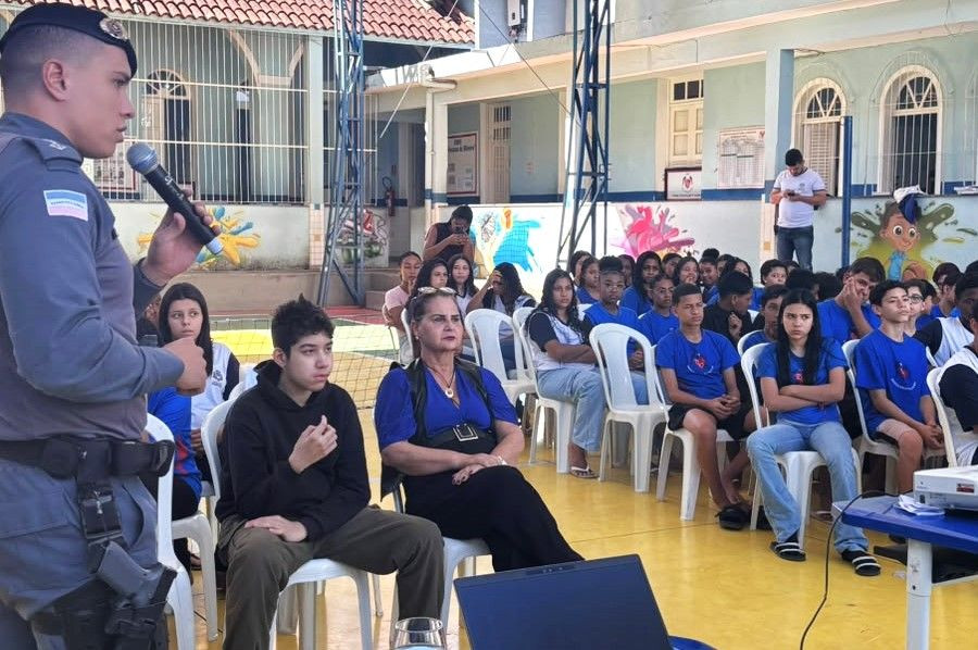 Policial militar em pé segurando um microfone enquanto fala para uma plateia de estudantes e profissionais da educação sentados na quadra poliesportiva de uma escola em Guaçuí.
