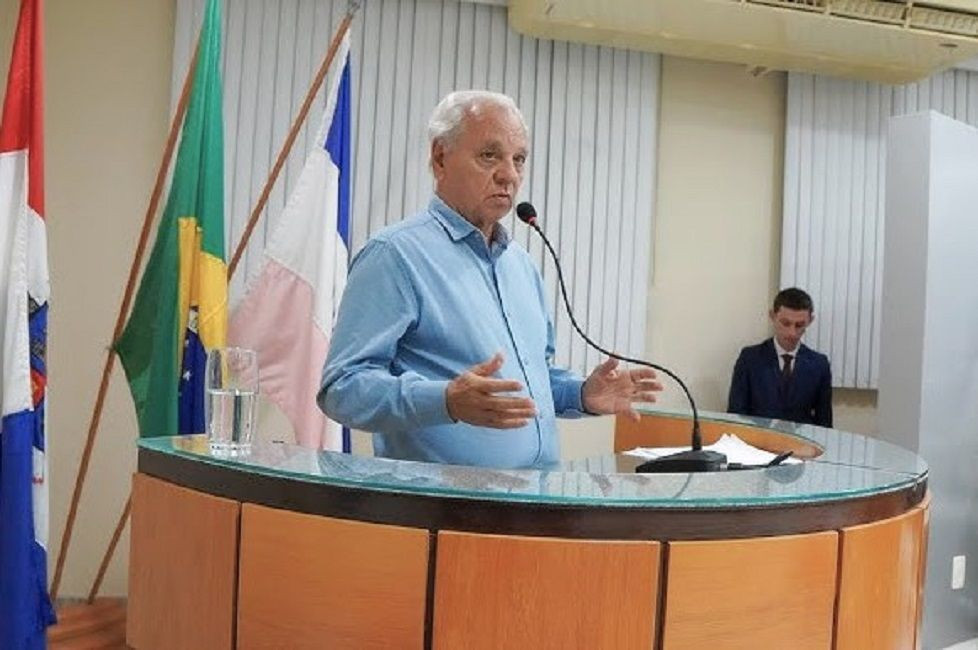 O candidato estadual Zé Ramos vestindo camisa social azul clara, em pé atrás de uma tribuna de madeira e vidro. Ele está discursando com as mãos abertas e em frente a um microfone de haste flexível. Ao fundo, veem-se as bandeiras do Brasil e do Estado do Espírito Santo, além de um homem de terno escuro em segundo plano.