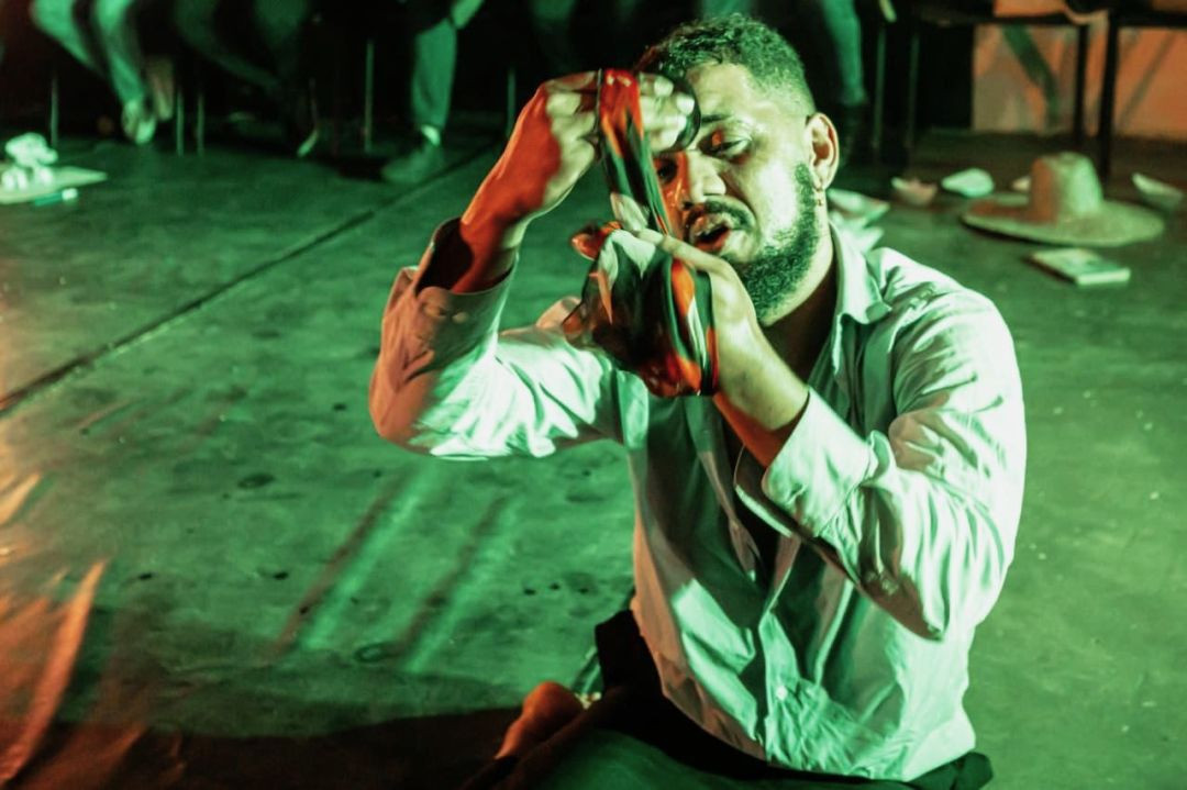 Ator ajoelhado sob iluminação verde, segurando um tecido colorido próximo ao rosto durante apresentação teatral.