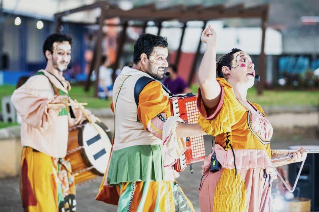 Três atores em trajes coloridos e maquiagem cênica apresentando-se ao ar livre; um toca sanfona e outro segura um instrumento de percussão.