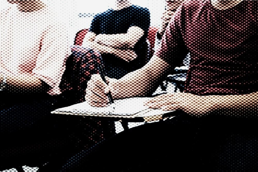 Estudante sentado à mesa escreve em um documento durante atividade acadêmica, representando a concessão de auxílio financeiro para estudantes de cursos superiores e técnicos.