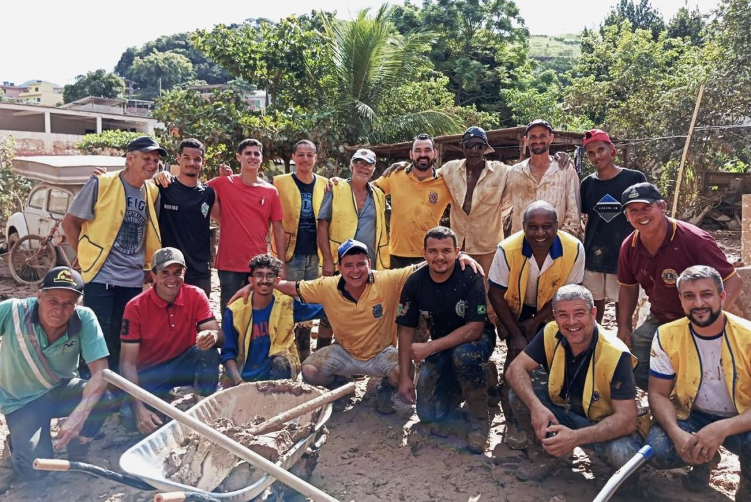 Fotografia colorida em ambiente externo. Um grupo de cerca de 15 homens, alguns vestindo coletes amarelos com o emblema do Lions e outros com roupas de trabalho sujas de terra, posa sorrindo. À frente, há um carrinho de mão. O fundo é composto por vegetação e construções simples.
