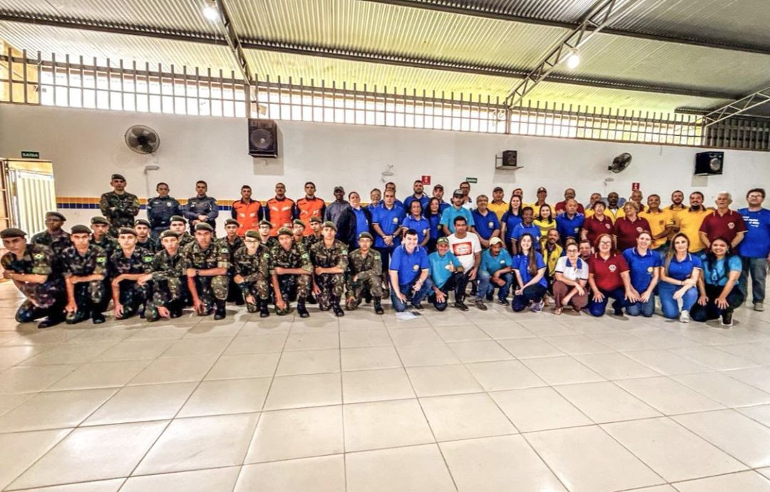 Uma foto de grupo ampla, tirada dentro de um salão de eventos com piso de cerâmica clara e teto de estrutura metálica. No centro, dezenas de pessoas estão de pé e ajoelhadas, posando para a foto. Elas vestem uniformes variados: alguns com camisas azuis ou amarelas do Lions, outros em fardas militares verdes, uniformes de bombeiro laranjas e camisas sociais. O salão tem paredes brancas e ventiladores.