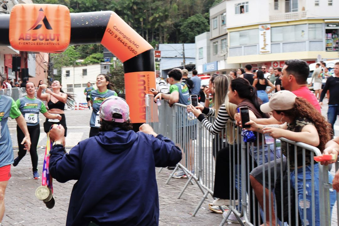 Fotografia de rua mostrando corredores cruzando a linha de chegada sob um pórtico inflável preto e laranja da Absolut Eventos Esportivos. Ao lado direito, o público observa o evento atrás de grades de proteção. Em primeiro plano, uma pessoa de costas segura medalhas penduradas. Ao fundo, veem-se prédios comerciais e o comércio local de Dores do Rio Preto.