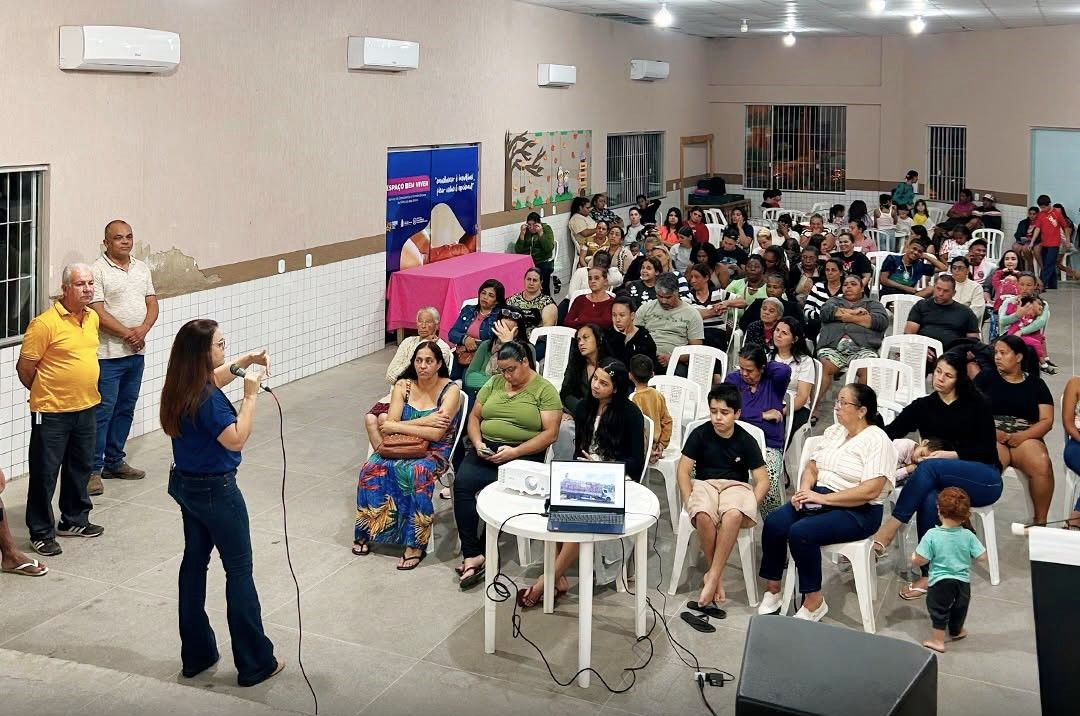 Uma mulher em pé, utilizando microfone e camiseta azul, fala para uma plateia de moradores sentados em cadeiras brancas durante palestra em um auditório.