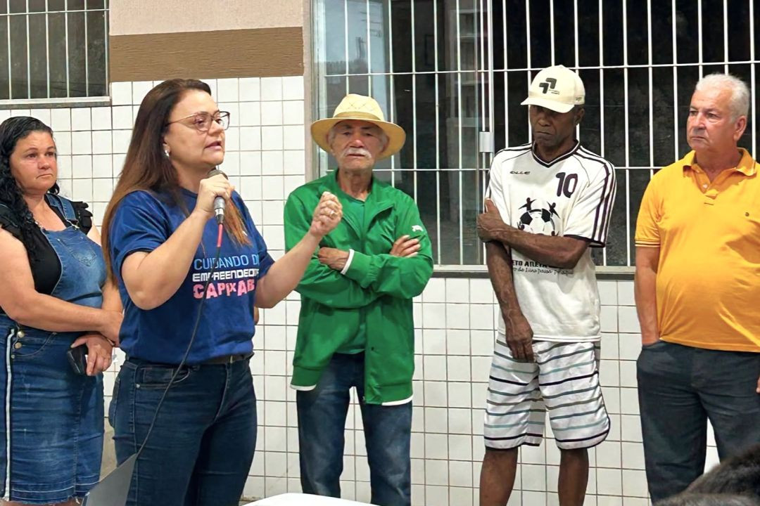 Mulher de óculos e camiseta azul discursa com microfone em mãos ao lado de quatro homens e uma mulher alinhados em pé perto de uma parede de azulejos claros.