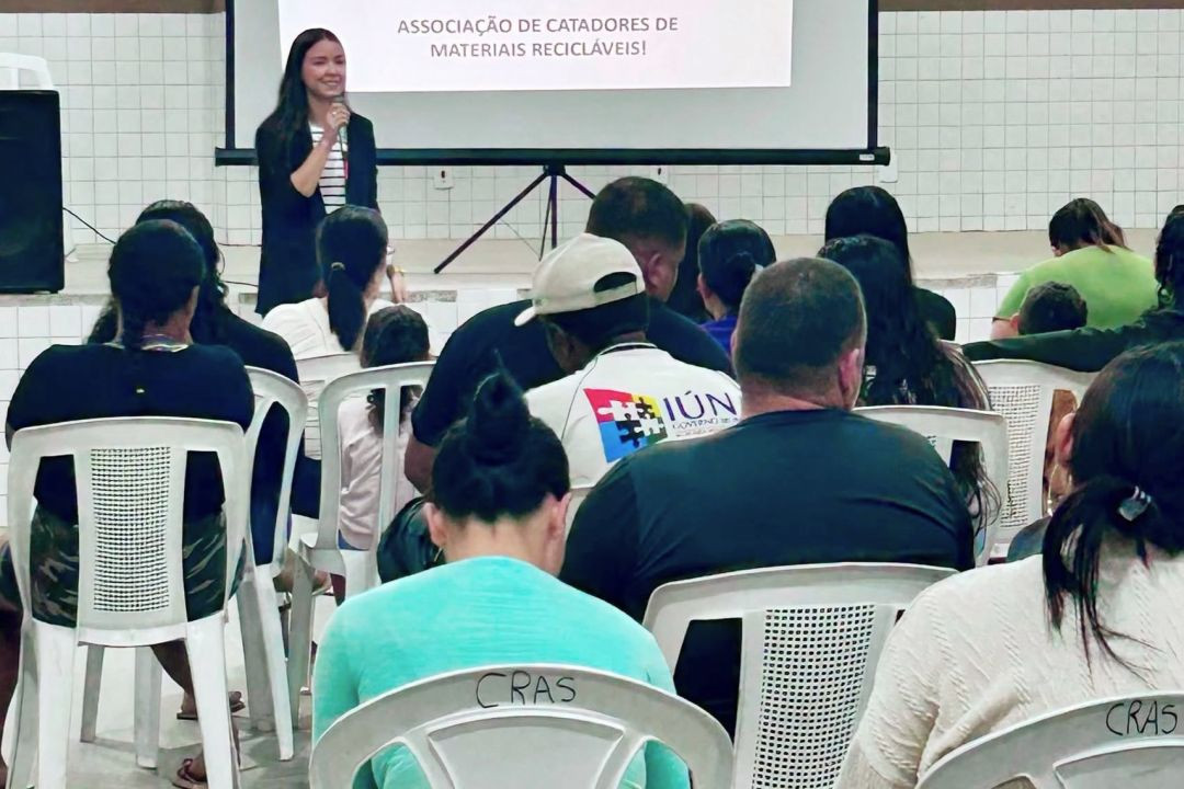Vista posterior de um auditório com pessoas sentadas acompanhando a apresentação de uma palestrante ao fundo, ao lado de um telão com a frase 