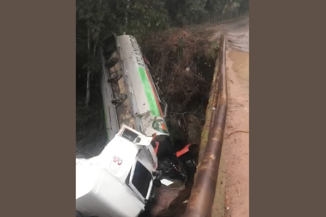 Fotografia na vertical mostrando um caminhão-tanque de cabine branca e tanque prateado tombado de lado no barranco de um córrego, ao lado de uma ponte com proteção metálica danificada em uma estrada de terra.