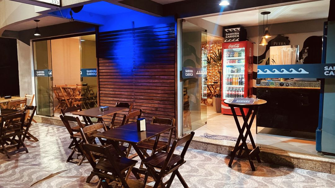 Fachada e área externa de uma lanchonete à noite, mostrando portas de vidro de correr, mesas na calçada e balcão interno iluminado.